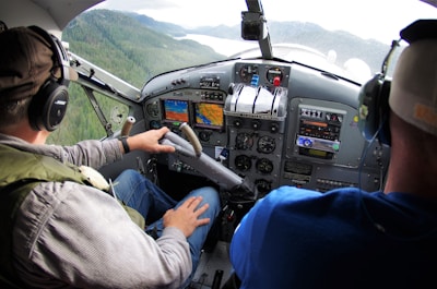 Two people are seated in the cockpit of a small aircraft, wearing headsets. The person on the left is operating the controls while the other observes. The cockpit features various instruments, dials, and screens. A view of forested mountains and a body of water is visible through the windshield.