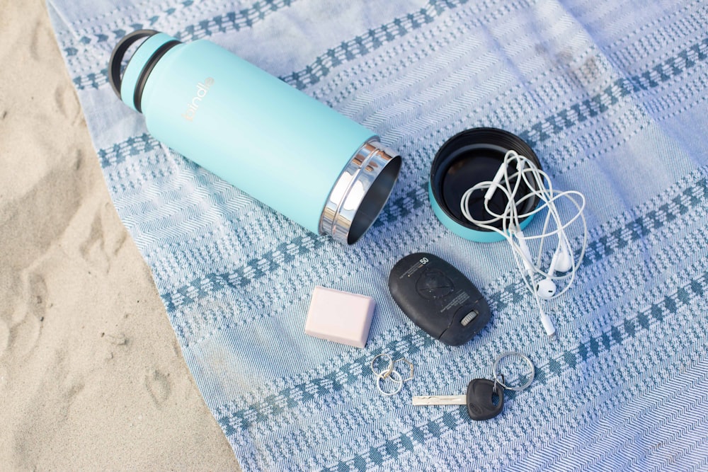 teal stainless steel travel mug with fob, key and earbuds on blue textile
