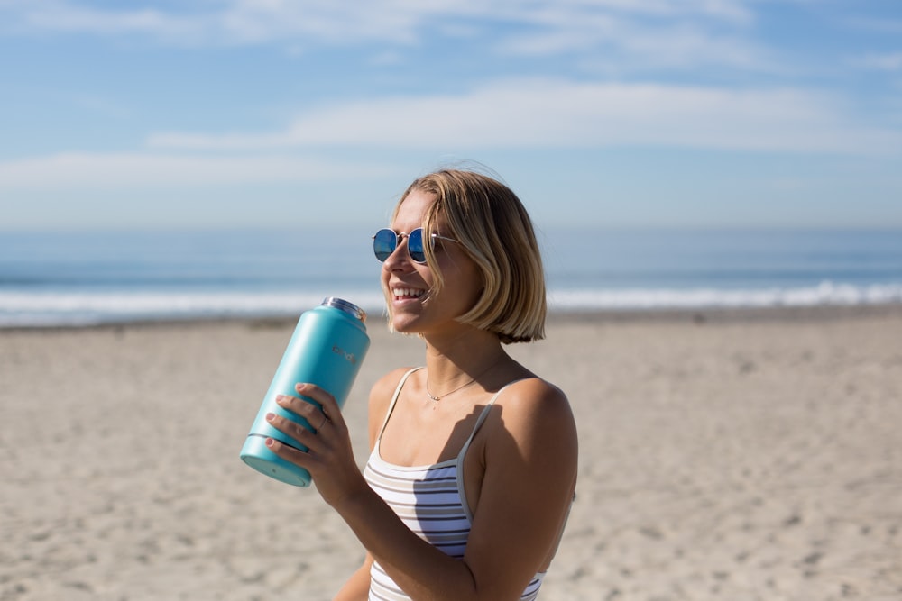 woman in white and brown pinstriped sleeveless top holding blue tumbler while smiling and standing near seashore at daytime