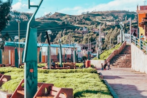 A vibrant park area with lush green grass and modern architectural elements. People are seen walking up the stone stairs and children are playing on the pathway. The background features a scenic view of rolling hills and dense trees. The park is equipped with uniquely designed lamp posts and benches.