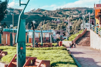 A vibrant park area with lush green grass and modern architectural elements. People are seen walking up the stone stairs and children are playing on the pathway. The background features a scenic view of rolling hills and dense trees. The park is equipped with uniquely designed lamp posts and benches.
