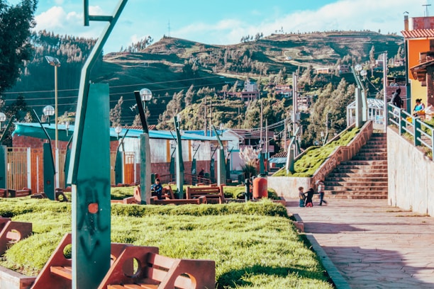 A vibrant park area with lush green grass and modern architectural elements. People are seen walking up the stone stairs and children are playing on the pathway. The background features a scenic view of rolling hills and dense trees. The park is equipped with uniquely designed lamp posts and benches.