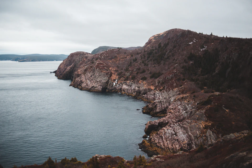 The image depicts a serene coastal landscape with a rocky cliff face jutting out into the ocean, its surface reflecting the overcast sky above. The cliff is covered in patches of vegetation and lichen, adding a touch of green to the otherwise brown and gray palette. The water below mirrors the cloudy sky, creating a mirror image that enhances the sense of depth and perspective. In the distance, beyond the rocky cliff face, there are hills rising up against the horizon, providing a sense of scale and grandeur to the scene.