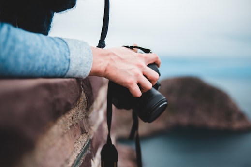 A person's hand holds a DSLR camera over a stone wall, above a blurred landscape with an out-of-focus view of nature and distant hills. The image conveys a sense of exploration and photography.