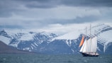 A sailing ship with large white sails and an orange detail on one sail is navigating through a body of water with a backdrop of snow-capped mountains. The sky is overcast with a layer of gray clouds, adding to the serene and slightly somber atmosphere.