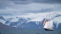 A sailing ship with large white sails and an orange detail on one sail is navigating through a body of water with a backdrop of snow-capped mountains. The sky is overcast with a layer of gray clouds, adding to the serene and slightly somber atmosphere.
