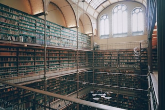 An elaborate and spacious multilevel library with ornate metal railings and countless rows of bookshelves filled with books. Large, arched windows allow natural light to illuminate the interior, creating a serene and studious atmosphere. A spiral staircase adds to the architectural beauty, while the overall design suggests a historic or classic feel.