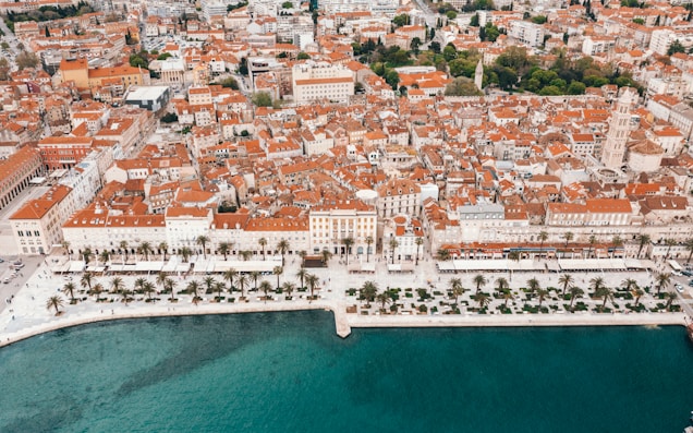 An aerial view of a coastal city featuring terracotta rooftops and historic architecture. The waterfront is lined with palm trees and a promenade, adjacent to clear turquoise water. Urban sprawl is evident as it extends beyond the shoreline, incorporating green spaces and dense residential and commercial buildings.