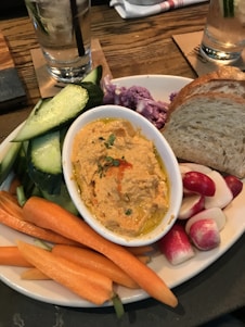 An appetizing plate featuring a variety of fresh vegetables including slices of cucumber, carrot sticks, radishes, and purple cauliflower. A small dish of hummus garnished with herbs is placed alongside slices of rustic bread. Two glasses of water with ice and cucumber slices are set on the wooden table.