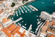 An aerial view of a historic coastal city featuring a bustling harbor filled with numerous small boats. The image shows the city's ancient stone walls and buildings with red-tiled roofs extending around the clear blue water of the bay.