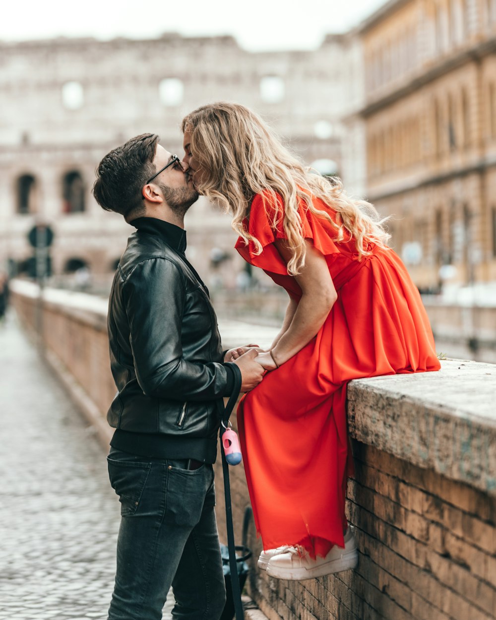 man standing and kissing woman sitting on concrete wall