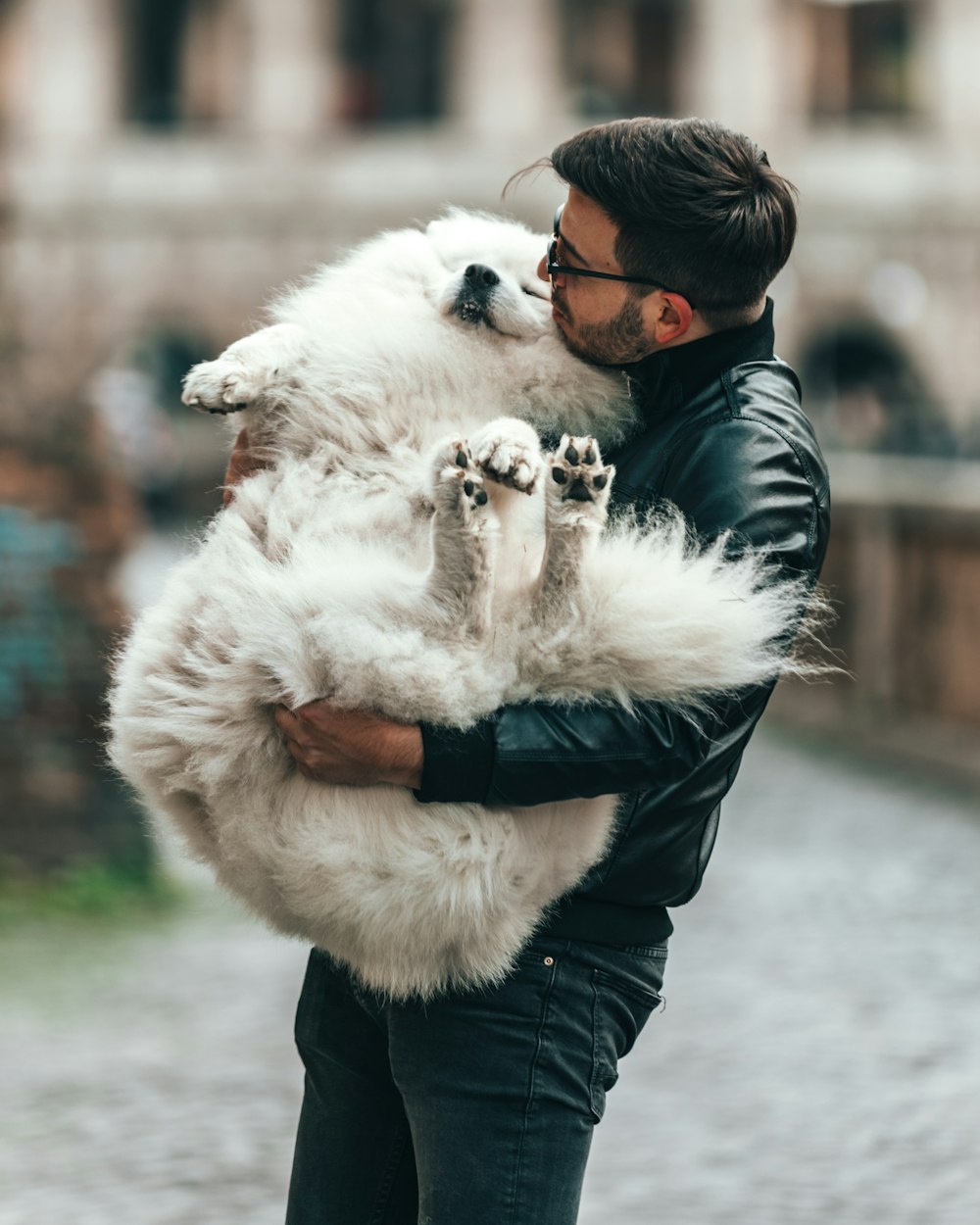 man in eyeglasses and black jacket carrying white dog