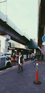 Pedestrians walk along a street, with a modern bus visible on the road and an elevated train track running above. A traffic warden vehicle is parked nearby, and a person is crossing in front of the bus. The scene is urban and suggests a busy city environment.