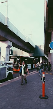 Pedestrians walk along a street, with a modern bus visible on the road and an elevated train track running above. A traffic warden vehicle is parked nearby, and a person is crossing in front of the bus. The scene is urban and suggests a busy city environment.
