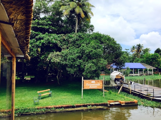 A serene riverside setting with lush greenery, a palm tree, and a sign for Punnamada Resort. A small wooden bench sits on the grassy bank by the water. A building with a blue roof and a covered boat are visible across a wooden dock.