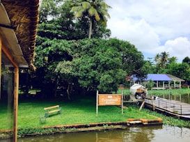 A serene riverside setting with lush greenery, a palm tree, and a sign for Punnamada Resort. A small wooden bench sits on the grassy bank by the water. A building with a blue roof and a covered boat are visible across a wooden dock.