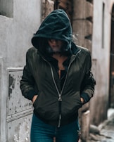 A person wearing a dark hoodie and a bomber jacket stands in an urban setting. The hood partly conceals their face, and their hands are in the jacket pockets. Smoke or mist appears around them, possibly exhaled, adding a mysterious or contemplative atmosphere. The surroundings include aged walls and a narrow alley.