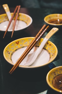 A table setting featuring Asian-style bowls with yellow and ornate patterns, complemented by wooden chopsticks and white porcelain spoons. The background includes additional matching bowls, creating a cohesive and inviting dining atmosphere.