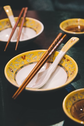 A table setting featuring Asian-style bowls with yellow and ornate patterns, complemented by wooden chopsticks and white porcelain spoons. The background includes additional matching bowls, creating a cohesive and inviting dining atmosphere.