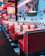 A retro-style diner with red and white upholstered booths arranged along the wall. Metal pendant lights hang from the ceiling above each booth. The walls are adorned with vintage car hubcaps and license plates. A checkered black-and-white floor enhances the classic diner aesthetic. There are menus and small condiment holders on the tables.