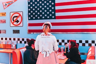 A retro diner setting with a large American flag on the wall. A waitress wearing a 1950s-inspired uniform is standing near a booth where two people are seated, engaged in conversation. The decor includes vintage signage, including a no U-turn sign and various license plates. Classic diner booths with red and white upholstery are visible.