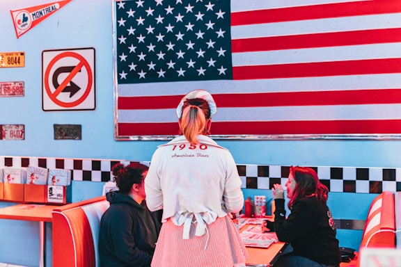 A retro diner setting with a large American flag on the wall. A waitress wearing a 1950s-inspired uniform is standing near a booth where two people are seated, engaged in conversation. The decor includes vintage signage, including a no U-turn sign and various license plates. Classic diner booths with red and white upholstery are visible.