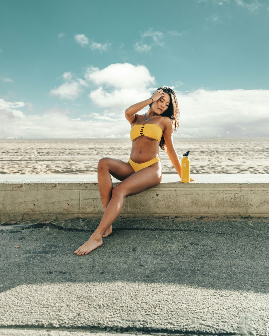 woman wearing yellow bikini set sitting on concrete with yellow bottle in front of sea