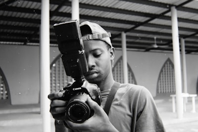 A person is holding a professional DSLR camera with a large external flash unit attached. They appear to be looking intently at the camera's display or viewfinder, possibly checking a photo they just took or adjusting settings. The background features architectural structures with geometric patterns and columns.