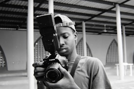 A person is holding a professional DSLR camera with a large external flash unit attached. They appear to be looking intently at the camera's display or viewfinder, possibly checking a photo they just took or adjusting settings. The background features architectural structures with geometric patterns and columns.