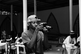 A person is engrossed in taking a photo with a professional camera, wearing a baseball cap backward and a casual t-shirt. The background features a group of people seated at tables with drinks, suggesting a social gathering or event, set in a covered outdoor area with decorative archways.