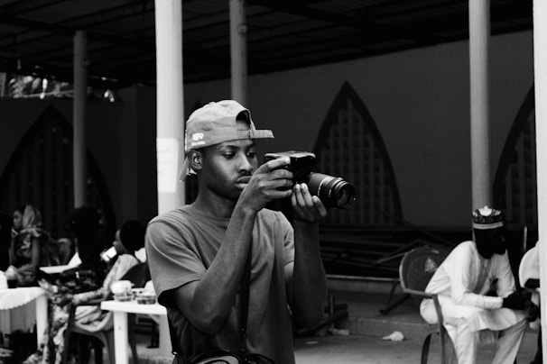 A person is engrossed in taking a photo with a professional camera, wearing a baseball cap backward and a casual t-shirt. The background features a group of people seated at tables with drinks, suggesting a social gathering or event, set in a covered outdoor area with decorative archways.