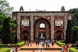 A historical building with large arched doorways and windows, constructed from stone and red sandstone. It features symmetrical patterns and design elements typical of Mughal architecture. Well-maintained gardens with trimmed hedges and trees line the pathway leading to the entrance. A variety of people are walking along the path, some in colorful traditional attire.