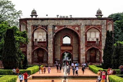 A historical building with large arched doorways and windows, constructed from stone and red sandstone. It features symmetrical patterns and design elements typical of Mughal architecture. Well-maintained gardens with trimmed hedges and trees line the pathway leading to the entrance. A variety of people are walking along the path, some in colorful traditional attire.