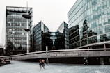 Tall, modern glass buildings with sleek, reflective surfaces stand alongside a wide pedestrian walkway. A few people are walking along, and there is a notable structure with the words 'more london riverside' displayed prominently. The sky is overcast, casting a muted tone over the scene.
