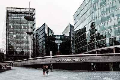 Tall, modern glass buildings with sleek, reflective surfaces stand alongside a wide pedestrian walkway. A few people are walking along, and there is a notable structure with the words 'more london riverside' displayed prominently. The sky is overcast, casting a muted tone over the scene.