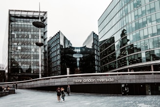Tall, modern glass buildings with sleek, reflective surfaces stand alongside a wide pedestrian walkway. A few people are walking along, and there is a notable structure with the words 'more london riverside' displayed prominently. The sky is overcast, casting a muted tone over the scene.