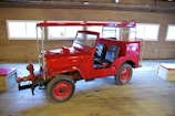 A bright red vintage firefighting vehicle with an open cabin is parked inside a wooden building. The vehicle has large tires and is equipped with firefighting equipment on the front. The wooden interior of the building features several rectangular windows, allowing natural light to illuminate the scene.