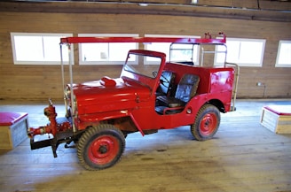 A bright red vintage firefighting vehicle with an open cabin is parked inside a wooden building. The vehicle has large tires and is equipped with firefighting equipment on the front. The wooden interior of the building features several rectangular windows, allowing natural light to illuminate the scene.