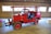 A bright red vintage firefighting vehicle with an open cabin is parked inside a wooden building. The vehicle has large tires and is equipped with firefighting equipment on the front. The wooden interior of the building features several rectangular windows, allowing natural light to illuminate the scene.
