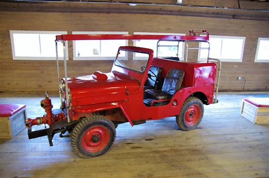 A bright red vintage firefighting vehicle with an open cabin is parked inside a wooden building. The vehicle has large tires and is equipped with firefighting equipment on the front. The wooden interior of the building features several rectangular windows, allowing natural light to illuminate the scene.