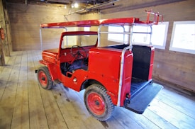 A vintage red jeep with a metal frame on top is parked inside a spacious wooden room. The jeep, with an open top design, is positioned on wooden floorboards, and there are large windows on the right side letting in natural light.