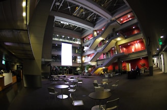 A spacious, modern atrium in an office building with multiple levels of walkways and staircases visible. The area features contemporary furniture including tables and chairs in the foreground, while the background is lined with red accent walls and illuminated by spotlights. Several plants are positioned throughout, and a large, bright screen is prominently displayed on one of the walls, contributing to the ambient light in the area.
