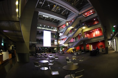 A spacious, modern atrium in an office building with multiple levels of walkways and staircases visible. The area features contemporary furniture including tables and chairs in the foreground, while the background is lined with red accent walls and illuminated by spotlights. Several plants are positioned throughout, and a large, bright screen is prominently displayed on one of the walls, contributing to the ambient light in the area.