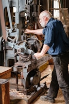 An older man with a white beard and glasses is operating a large, vintage industrial machine in a workshop setting. The machine is made of metal with visible gears and levers. Wood shavings cover the floor, suggesting woodworking or machining activity. He is focused on adjusting the machinery, dressed in a dark blue shirt and black pants, conveying a work-focused environment.