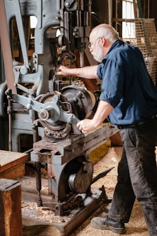 An older man with a white beard and glasses is operating a large, vintage industrial machine in a workshop setting. The machine is made of metal with visible gears and levers. Wood shavings cover the floor, suggesting woodworking or machining activity. He is focused on adjusting the machinery, dressed in a dark blue shirt and black pants, conveying a work-focused environment.