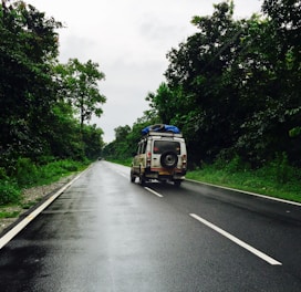 A vehicle with a roof rack and a spare tire drives down a wet, asphalt road surrounded by lush green trees. The sky is overcast, suggesting recent or impending rain. The scene conveys a sense of travel or adventure, capturing a moment of a road trip in a natural setting.