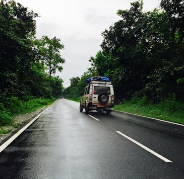 A vehicle with a roof rack and a spare tire drives down a wet, asphalt road surrounded by lush green trees. The sky is overcast, suggesting recent or impending rain. The scene conveys a sense of travel or adventure, capturing a moment of a road trip in a natural setting.