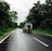 A vehicle with a roof rack and a spare tire drives down a wet, asphalt road surrounded by lush green trees. The sky is overcast, suggesting recent or impending rain. The scene conveys a sense of travel or adventure, capturing a moment of a road trip in a natural setting.