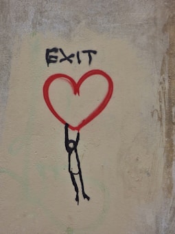 Street art on a wall featuring a black stick figure hanging from a large red heart. Above the heart, the word 'EXIT' is painted in black.