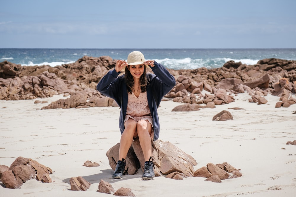 woman in blue jacket sitting on rock by the beach while holding sun hat